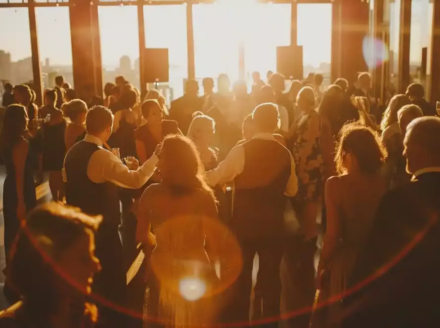 Guests mingle and dance at a lively wedding reception, with the city skyline visible through the windows.